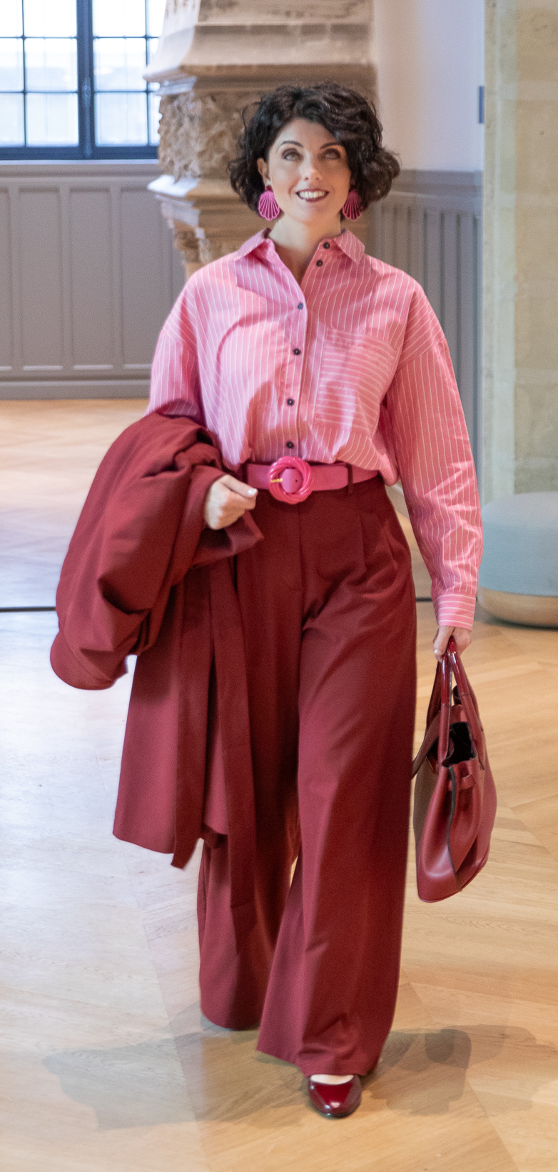 Luisa Fenu, debout en pied dans un hall en pierre, manteau bordeaux à la main